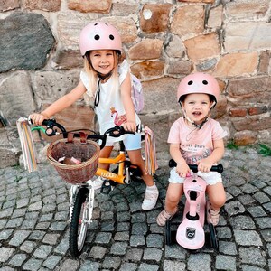 May include: Two children wearing pink helmets ride toy vehicles on a brick pathway. The girl on the left rides a yellow bicycle with a wicker basket and colourful streamers. The girl on the right rides a pink scooter with a white handlebar.