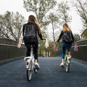 May include: Two people riding bicycles down a paved path on a bridge. Both bicycles have streamers attached to the handlebars. The person in the front is wearing a black leather backpack with fringe. The person in the back is wearing a black backpack with a drawstring closure.