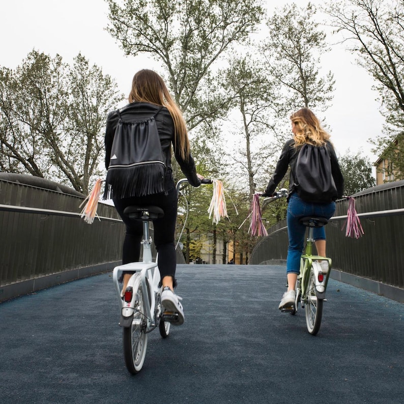 May include: Two people riding bicycles down a paved path on a bridge. Both bicycles have streamers attached to the handlebars. The person in the front is wearing a black leather backpack with fringe. The person in the back is wearing a black backpack with a drawstring closure.
