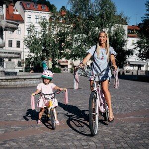 May include: A woman and a child riding bicycles with pink streamers attached to the handlebars. The woman is wearing a blue dress and the child is wearing a pink dress and a watermelon helmet.