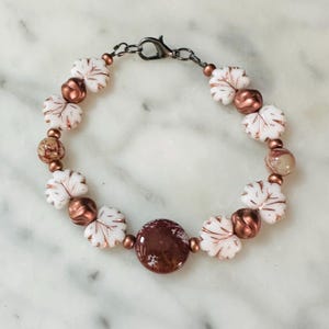 May include: A beaded bracelet featuring white leaf-shaped beads, copper-colored beads, and a large, round, speckled brown bead. The bracelet is strung on a dark metal chain with a lobster clasp.