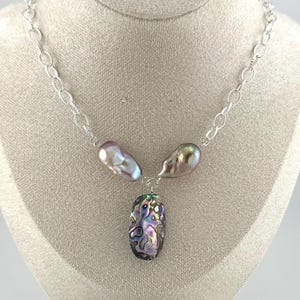May include: A silver chain necklace with three large, iridescent pearls. Two pearls are a light purple color, and the third is an oval-shaped abalone shell with a mix of colors. The necklace is displayed on a mannequin.