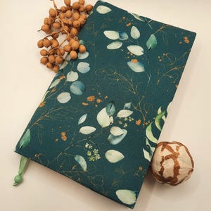 Könnte beinhalten: Ein tealblauer Notizbuchumschlag mit einem goldenen und weißen Blumenmuster. Der Umschlag hat ein grünes Band als Lesezeichen.