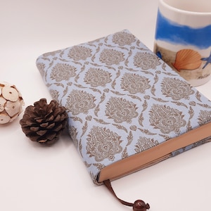 Könnte beinhalten: Ein Hardcover-Buch mit hellblauem Einband und braunem Paisley-Muster. Ein braunes Lesezeichen mit dekorativem Ende ist sichtbar. Eine dekorative Holzkugel und ein Tannenzapfen liegen neben dem Buch. Eine Tasse mit einer Strandszene befindet sich im Hintergrund.