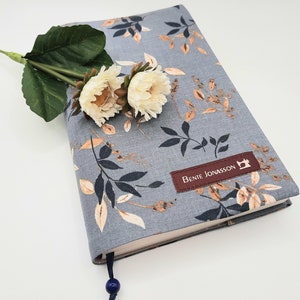 Könnte beinhalten: Ein grauer Stoffbuchumschlag mit einem floralen Muster in Braun- und Weißtönen. Der Umschlag hat ein braunes Etikett mit dem Text "Bente Jonasson" und einem Nähmaschinen-Symbol.