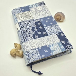 Könnte beinhalten: Ein Buchumschlag mit einem Patchwork-Design in Blau- und Weißtönen mit Paisley-Mustern. Ein blaues Lesezeichen mit einer Perle ist angebracht. Muscheln sind in der Nähe des Buches platziert.