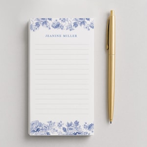 May include: A white notepad with blue floral designs at the top and bottom, and lined pages. The notepad has the name "JEANINE MILLER" printed in blue. A gold pen is positioned to the right of the notepad.