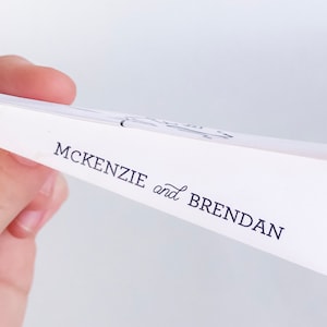 May include: White paper with the names "MCKENZIE and BRENDAN" printed in black script. The paper is held at an angle, revealing the text and a glimpse of a design at the top. The background is a soft, neutral color.
