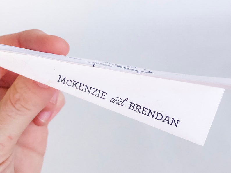 May include: White paper with the names "MCKENZIE and BRENDAN" printed in black script. The paper is held at an angle, revealing the text and a glimpse of a design at the top. The background is a soft, neutral color.