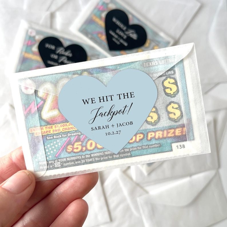 May include: A lottery ticket favor with a light blue heart-shaped sticker that reads "We Hit the Jackpot!" with names and a date. The ticket is inside a small, clear envelope. Other favors are visible in the background.