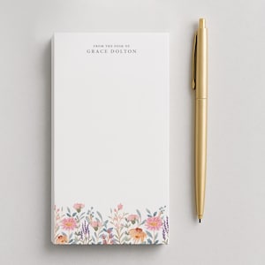 May include: A white notepad with a floral design at the bottom and the text "FROM THE DESK OF GRACE DOLTON" at the top. A gold pen is positioned to the right of the notepad.