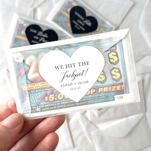 May include: Clear cellophane packets containing lottery tickets, each with a white heart-shaped sticker that reads "WE HIT THE Jackpot!" with names and a date. Additional black heart stickers are visible.