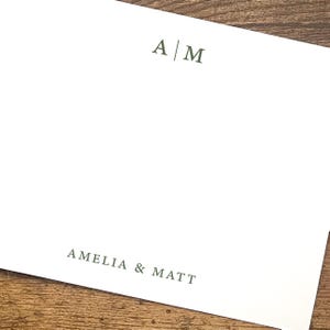 May include: A rectangular white stationery card featuring dark green text. The top center displays the initials A | M separated by a vertical line. The bottom center features the names AMELIA & MATT printed in a clean, serif font style.
