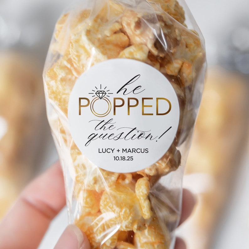 Pu&ograve; includere: Un sacchetto di bomboniere bianco pieno di popcorn e un adesivo rotondo che dice "Ha fatto la proposta!" con un anello d'oro e una grafica di diamante. L'adesivo include anche i nomi "Lucy + Marcus" e la data "10.18.25".
