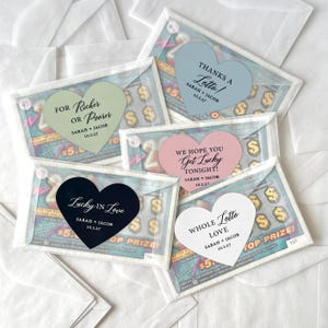 May include: A collection of lottery ticket favors with heart-shaped stickers. The stickers have phrases like "Lucky in Love" and "Thanks a Lotto!" with names and dates. The tickets are in clear envelopes.