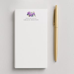 May include: A white notepad with the text "FROM THE DESK OF GRACE DOLTON" and a purple floral design. A gold pen is positioned vertically next to the notepad. The notepad and pen are on a white surface.