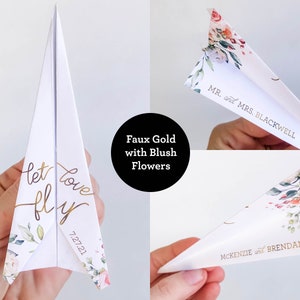 May include: White paper airplane with gold foil text that reads "let love fly" and "7.27.21". The airplane is decorated with blush-colored floral accents. The image also shows the names "Mr. and Mrs. Blackwell" and "McKenzie and Brendan".