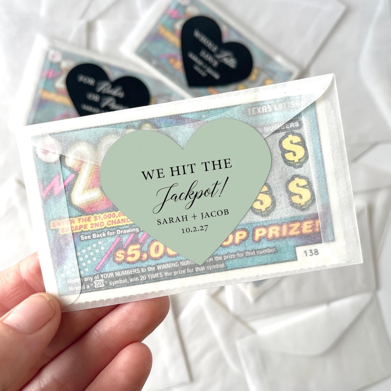 May include: A lottery ticket with a heart-shaped sticker that reads "We Hit the Jackpot!" with the names Sarah and Jacob and the date 10.2.27. The ticket is in a clear envelope, with other similar items in the background.