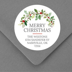 May include: A round white sticker with the words "MERRY CHRISTMAS" in a serif font, adorned with a watercolor illustration of green leaves and red berries. The sticker also includes an address in a serif font. The sticker is placed on a gray envelope.