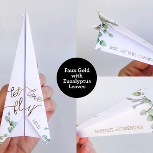 May include: White paper airplane with gold script reading "let love fly" and the date 7.27.21. The design includes eucalyptus leaves. Additional airplanes feature names like "Mr. and Mrs. Blackwell" and "McKenzie and Brendan".