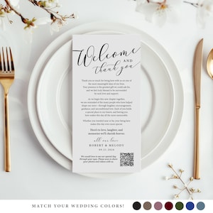 May include: A wedding reception place setting with a white card that reads "Welcome and Thank You" in elegant script. The card is on a white plate, with gold silverware and a sprig of flowers. The card includes a QR code.