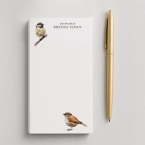 May include: A rectangular white notepad featuring a watercolor illustration of a small bird on a branch in the top left corner and another bird at the bottom, with the text from the desk of BRENDA SLOAN, accompanied by a gold metal pen.