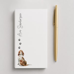 May include: A white notepad with the name "Ava Sanderson" in script, paw prints, and a watercolor illustration of a dog. A gold pen is to the right. The notepad is a pet-themed stationery item.