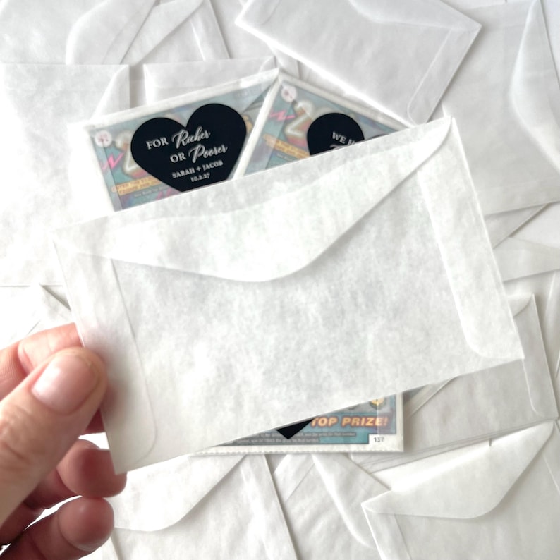 May include: A hand holding a small, white envelope with a scratch-off lottery ticket inside. The ticket has a black heart-shaped sticker that reads "For Richer or Poorer" and the names "Sarah + Jacob" with a date. Other envelopes are in the background.