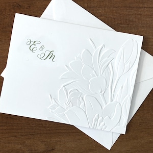 May include: A white rectangular card and matching envelope featuring a blind embossed floral design on the right side and the dark green printed monogram E & M in the upper left corner. The card has a smooth texture and clean edges.