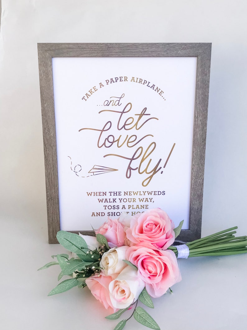 May include: A framed sign with the text "Take a paper airplane... and let love fly!" in gold script. A bouquet of pink and white roses and greenery rests in front of the frame. The frame is a light gray color.