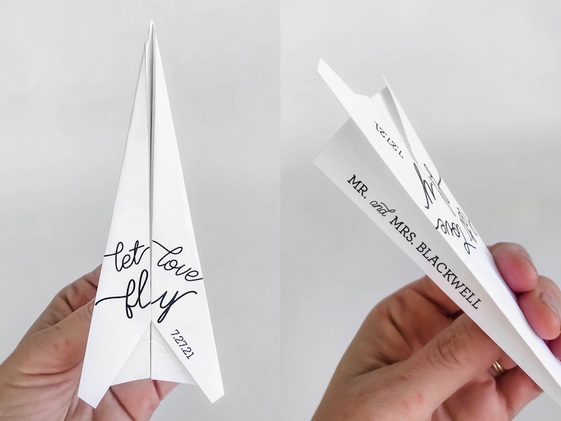 May include: White paper airplane with the words "let love fly" and the date "7.27.21" in black script. The airplane is folded and ready to launch. The other side reads "MR. and MRS. BLACKWELL".