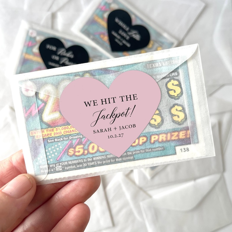 May include: A lottery ticket in a clear envelope, held by a hand. A pink heart-shaped sticker with the text "WE HIT THE Jackpot!" and the names "SARAH + JACOB" and the date "10.2.27" is on the front. Other tickets are in the background.