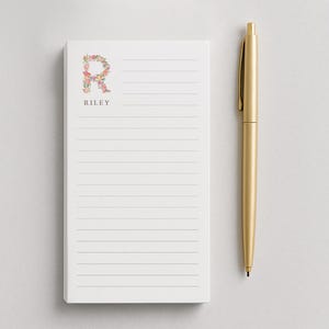May include: A white notepad with lined pages, featuring a floral letter "R" and the name "Riley" in a delicate font. A gold pen rests beside the notepad. The notepad is designed for writing notes.