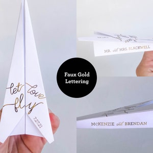 May include: White paper airplane with faux gold lettering that reads "let love fly" and the date "7.27.21". Additional airplanes feature names like "Mr. and Mrs. Blackwell" and "McKenzie and Brendan".
