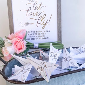 May include: A wedding decor display featuring paper airplanes in a galvanized metal tray. A sign with the words "...and let love fly!" is in the background. A bouquet of pink and white roses and greenery is on the left.