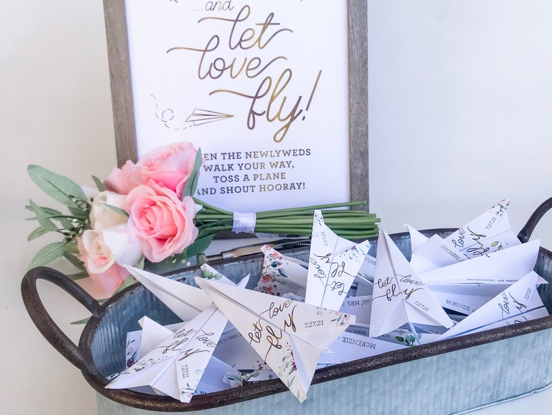 May include: A wedding decor display featuring paper airplanes in a galvanized metal tray. A sign with the words "...and let love fly!" is in the background. A bouquet of pink and white roses and greenery is on the left.