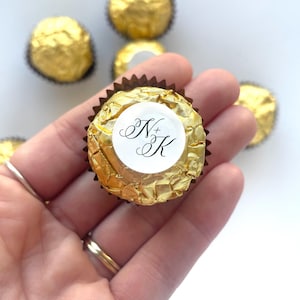 May include: A round chocolate candy wrapped in textured gold foil, featuring a white circular sticker on top with the black printed letters N+K in a decorative script font, surrounded by several other identical gold-wrapped candies.