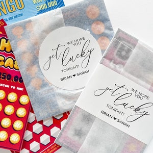 May include: A set of three white paper bags with a black and white sticker that says "get lucky tonight! Brian Sarah" and a black and white sticker that says "We hope you get lucky tonight! Brian Sarah". The bags contain scratch-off lottery tickets.