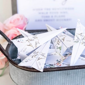 May include: A galvanized metal tray filled with white paper airplanes. Each plane has gold script that reads "let love fly" and the date "7.27.21". Floral accents and the names "Mr. and Mrs." are also visible. Pink flowers are in the background.