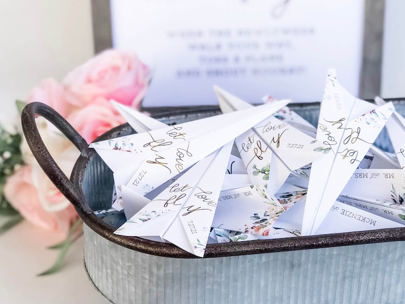 May include: A galvanized metal tray filled with white paper airplanes. Each plane has gold script that reads "let love fly" and the date "7.27.21". Floral accents and the names "Mr. and Mrs." are also visible. Pink flowers are in the background.