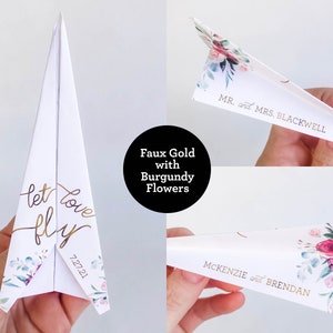May include: White paper airplane with gold lettering that reads "let love fly" and the date 7.27.21. The airplane has floral accents with burgundy flowers. The text "Faux Gold with Burgundy Flowers" is in a black circle. Other airplanes have names.