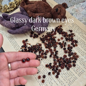 Könnte beinhalten: Eine Sammlung von dunkelbraunen, glänzenden Augen für Teddybären und andere Bastelarbeiten. Das Bild zeigt eine Vielzahl von Größen und Stilen, wobei einige Augen in einer Hand gehalten werden. Der Hintergrund umfasst Vintage-Papier und kleine Teddybärteile.
