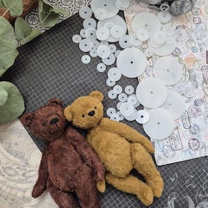 Op de afbeelding: Twee handgemaakte teddyberen, &eacute;&eacute;n bruin en &eacute;&eacute;n bruin, liggen samen op een donkergrijze ondergrond. Witte cirkelvormige componenten zijn eromheen verspreid, samen met papier met patronen en groen. De beren hebben gestikte details.