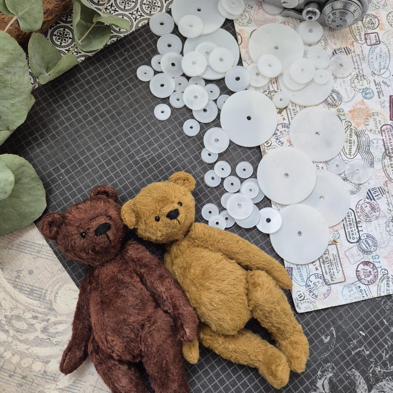 Op de afbeelding: Twee handgemaakte teddyberen, &eacute;&eacute;n bruin en &eacute;&eacute;n bruin, liggen samen op een donkergrijze ondergrond. Witte cirkelvormige componenten zijn eromheen verspreid, samen met papier met patronen en groen. De beren hebben gestikte details.