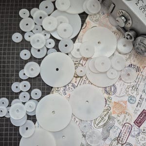 Op de afbeelding: Een assortiment witte, cirkelvormige horlogeonderdelen met centrale gaten, in verschillende maten, verspreid over een patroon. Een vintage camera is zichtbaar op de achtergrond, samen met postzegels en tekst.