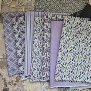 May include: A collection of seven folded fabric swatches in shades of purple and white. Patterns include a purple plaid, purple floral, purple stripes, purple socks, winter houses with trees, a purple polka dot, and birds on pine branches.