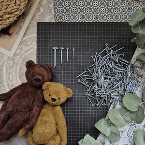 May include: Two plush teddy bears, one brown and one tan, sit next to each other. A pile of small nails and a few larger nails are on a black grid surface. Decorative paper and eucalyptus leaves are in the background.