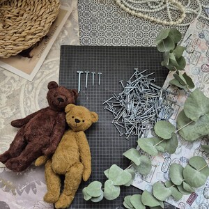 May include: Two handmade teddy bears, one brown and one tan, rest on a black grid surface. A pile of nails and a few individual nails are also present. The scene is decorated with eucalyptus leaves, a woven basket, and vintage-style paper.