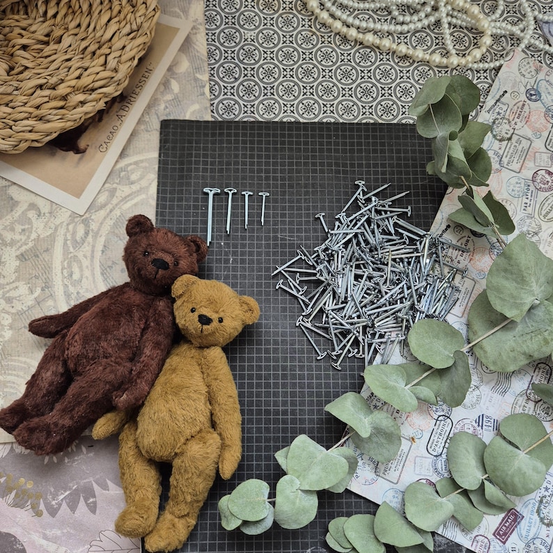 May include: Two handmade teddy bears, one brown and one tan, rest on a black grid surface. A pile of nails and a few individual nails are also present. The scene is decorated with eucalyptus leaves, a woven basket, and vintage-style paper.