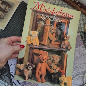 May include: A softcover book titled Miniaturbären machen mit Richard van Aalst featuring a cover illustration of several small, brown, tan, and golden plush teddy bears positioned around and inside a wooden cabinet with open doors.